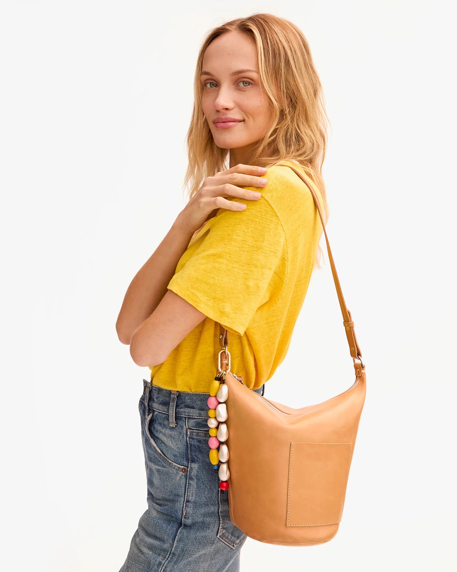 A woman in a yellow t-shirt and blue jeans stands sideways, smiling, with the Clare Vivier Petit Jeanne tan handwoven leather bucket bag, accented by a colorful beaded keychain accessory.