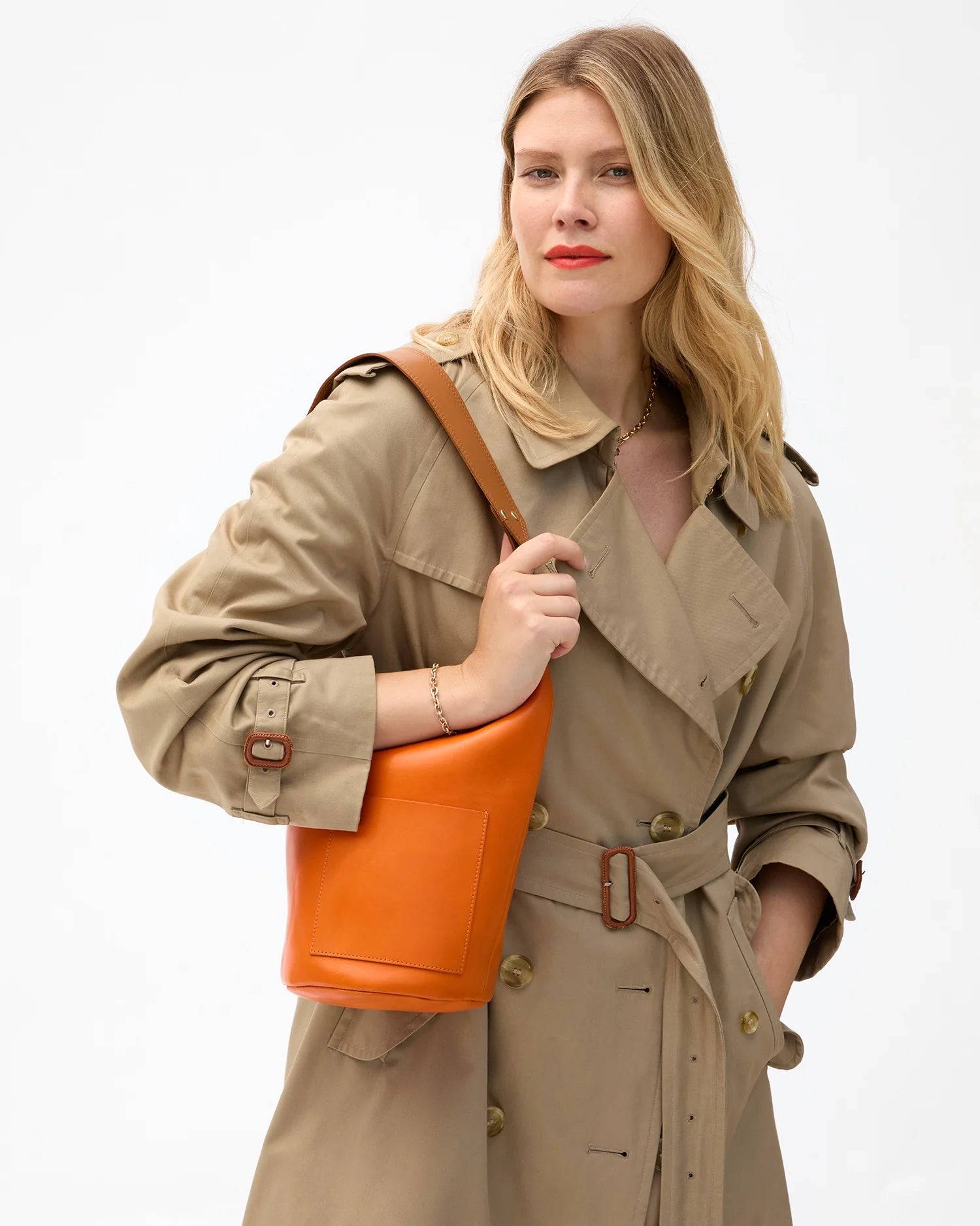 A woman in a beige trench coat holds the Clare Vivier Petite Jeanne Italian leather bucket bag, featuring a removable shoulder strap, and looks confidently at the camera against a plain white background.