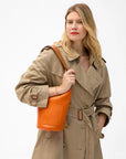 A woman in a beige trench coat holds the Clare Vivier Petite Jeanne Italian leather bucket bag, featuring a removable shoulder strap, and looks confidently at the camera against a plain white background.