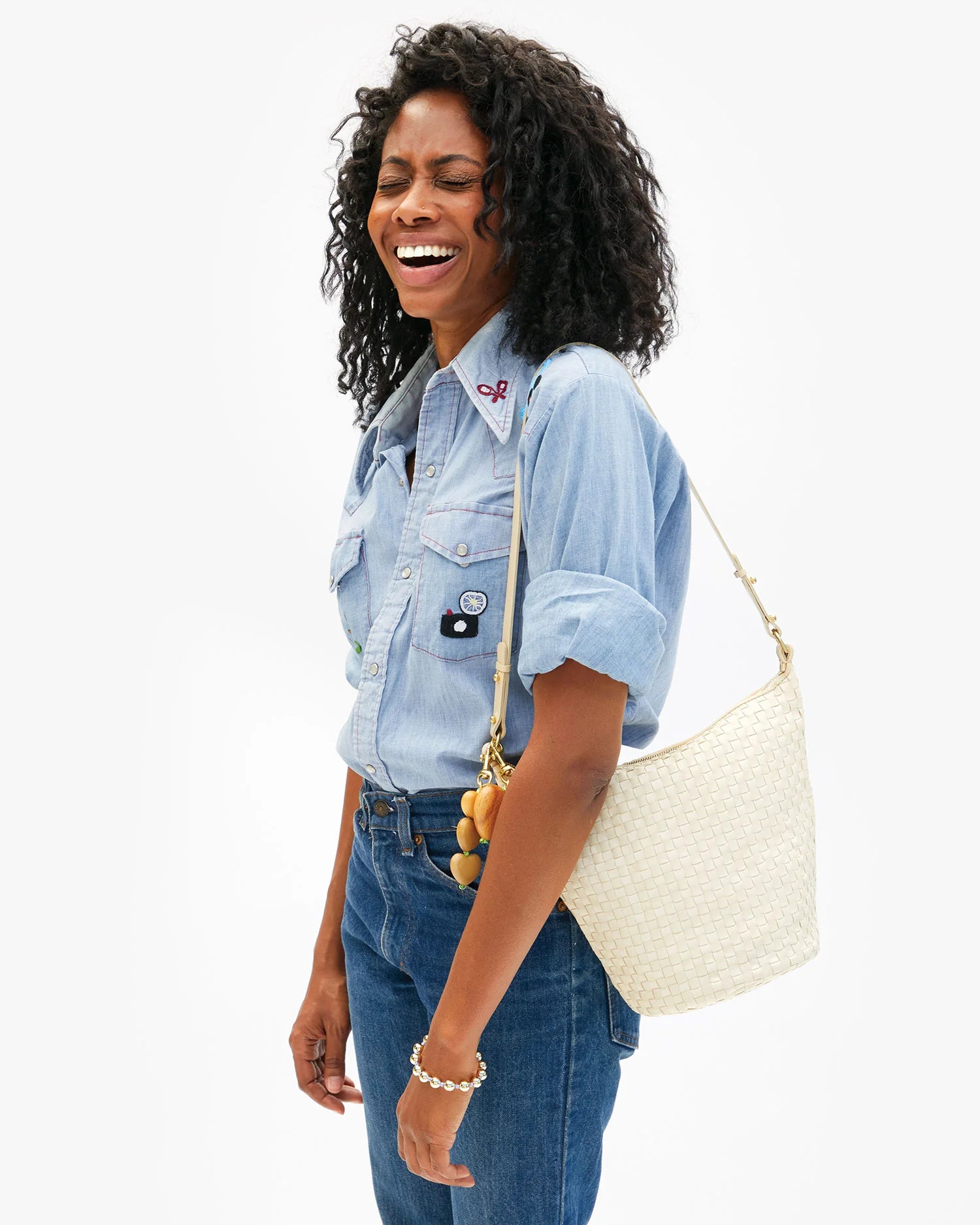 A woman with curly hair laughs while standing sideways, wearing a light denim shirt, blue jeans, and carrying the Clare Vivier Petit Jeanne handwoven leather bucket bag with removable shoulder strap against a plain white background.