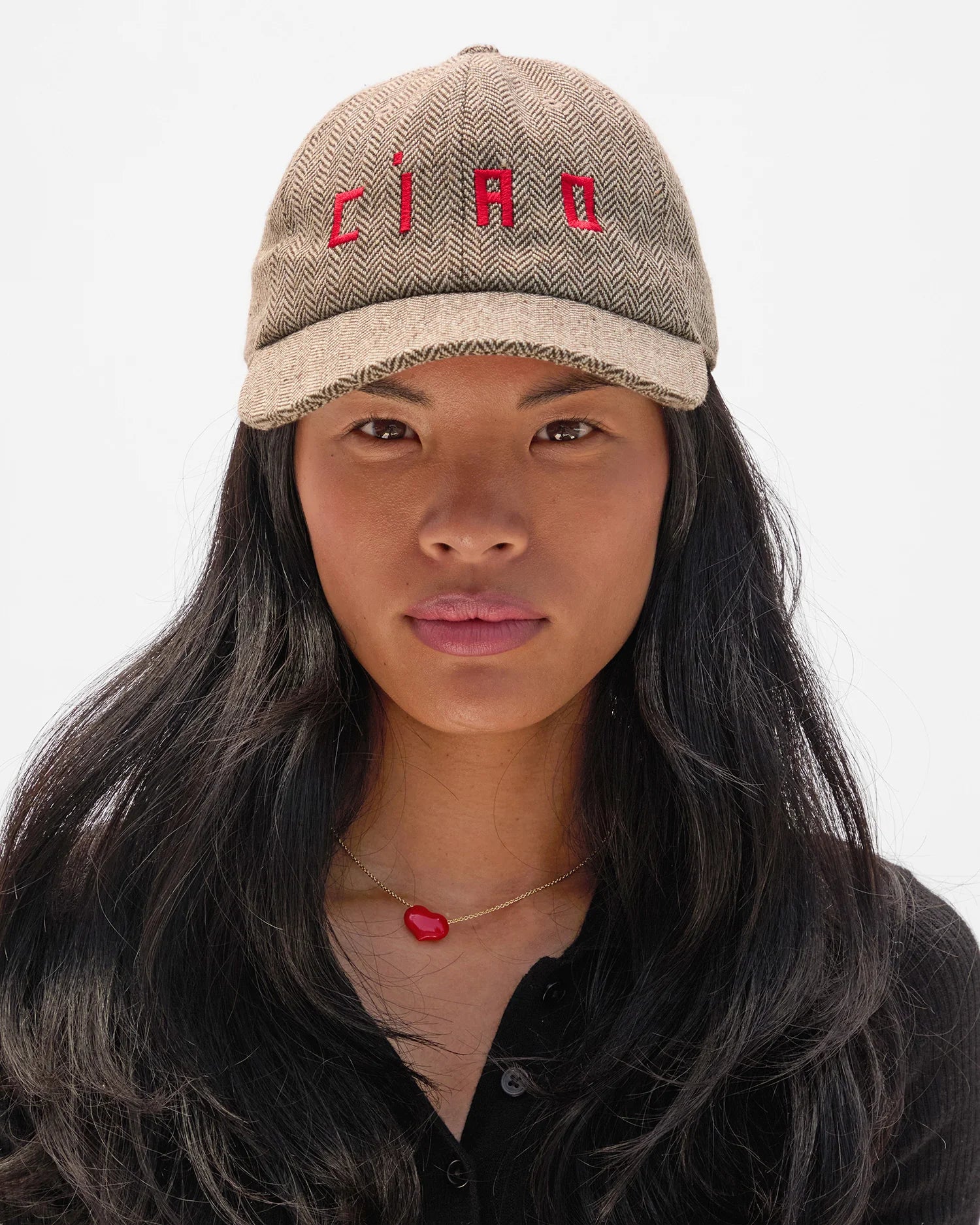 A woman with long black hair models the Clare Vivier Wool CIAO Baseball Hat, featuring tan herringbone wool embroidered with "CIAO" in red. She wears a black shirt and gold necklace with a red heart, facing forward against a plain white background.