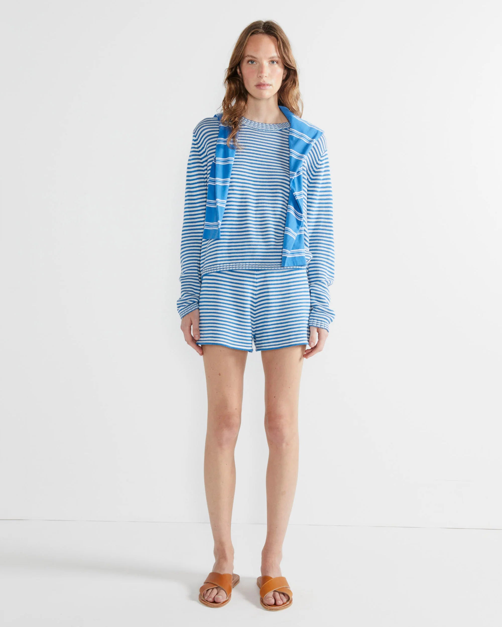 A woman stands against a white wall wearing The Peyton by Kule: a blue and white striped 100% cotton long-sleeve shirt, matching shorts, a striped knit sweater over her shoulders, and brown sandals.