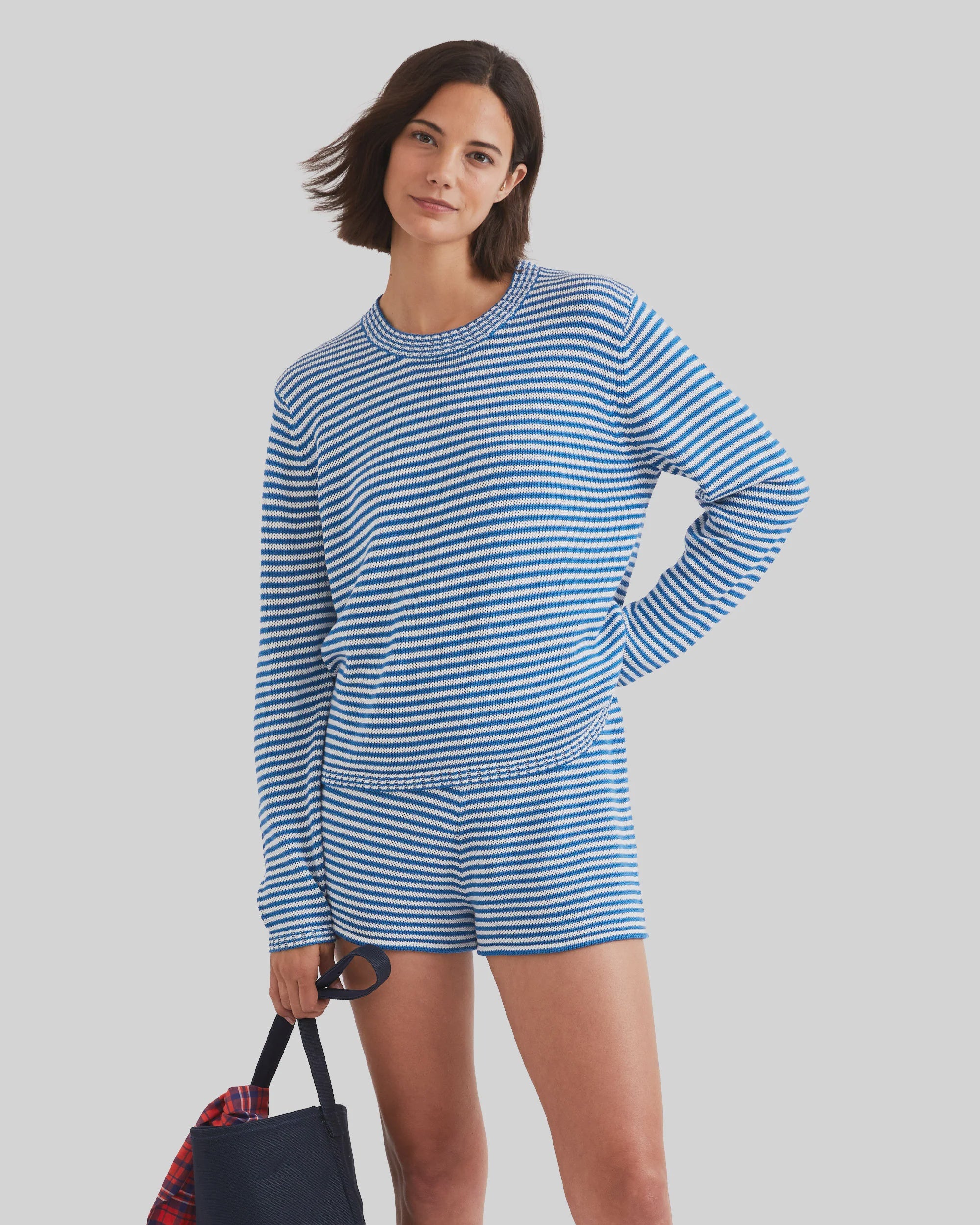 A woman with short brown hair smiles while wearing Kule’s The Peyton, a 100% cotton blue-and-white striped knit long-sleeve top and shorts, holding a navy tote with a red item inside against a light gray background.