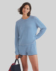 A woman with short brown hair smiles while wearing Kule’s The Peyton, a 100% cotton blue-and-white striped knit long-sleeve top and shorts, holding a navy tote with a red item inside against a light gray background.