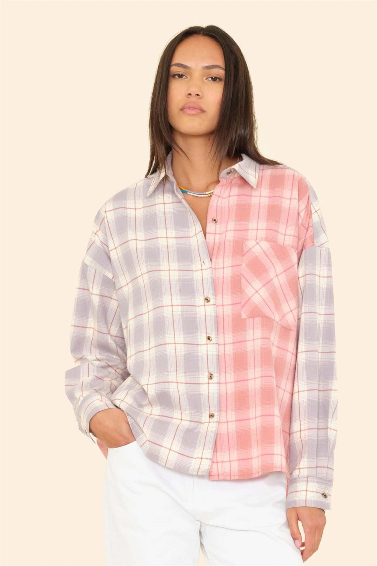 A woman with straight dark hair wears the Xirena Phillipa Shirt—a 100% cotton, collared, long-sleeve button-up in light purple and pink plaid—with white pants, standing against a plain beige background.