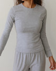 A woman wearing The Baby Rib Long Sleeve by Donni, along with matching pants, stands against a plain light-colored background. Her wavy hair cascades freely as she gazes to the side, one arm slightly bent. The subtle texture of the baby rib scallop fabric adds an elegant touch to her ensemble.