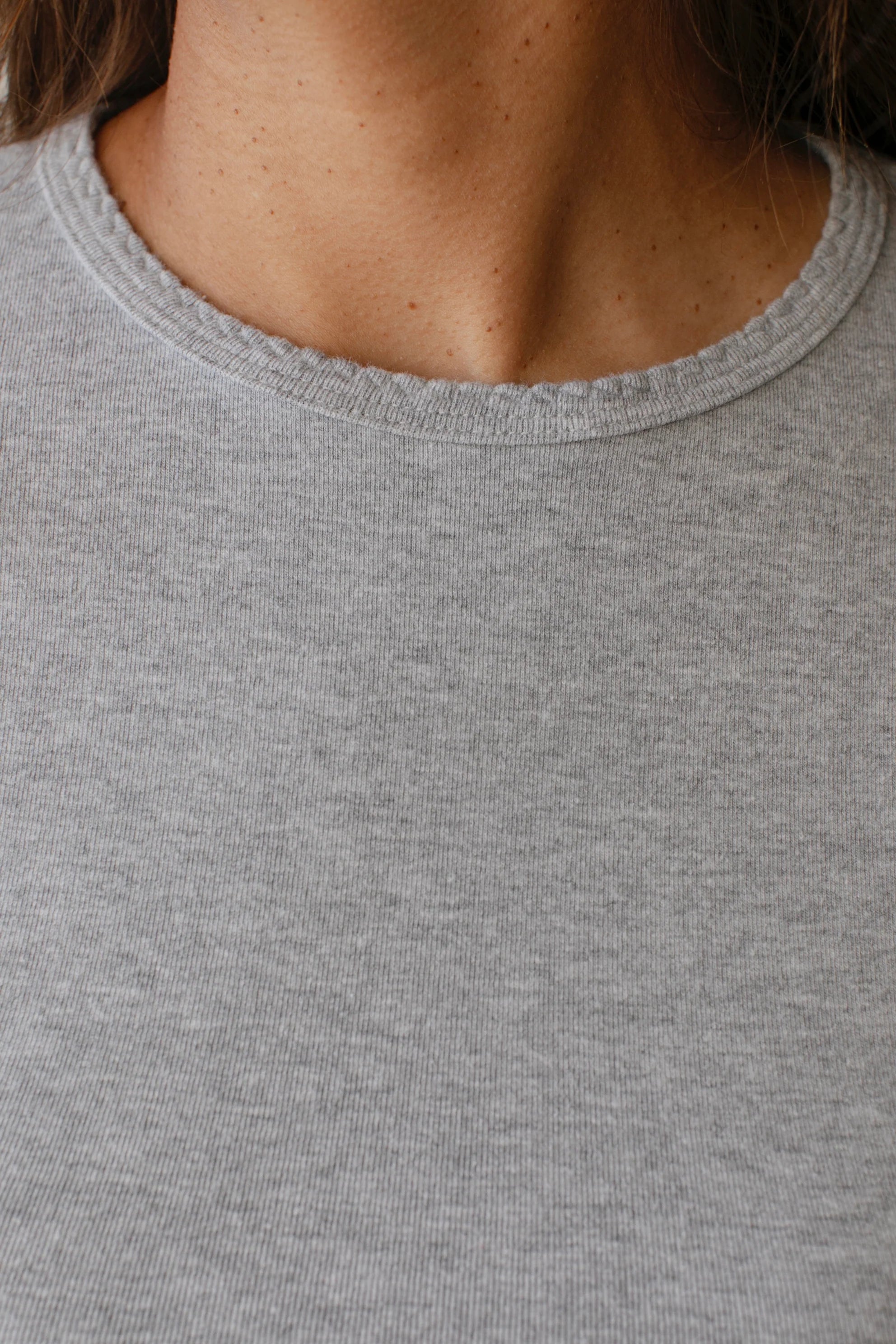 Close-up of a person wearing "The Baby Rib Long Sleeve" by Donni in grey. The image focuses on the neckline and upper chest area, highlighting the fabric's Baby Rib Scallop texture and complementing the skin's natural complexion.