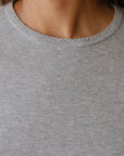 Close-up of a person wearing "The Baby Rib Long Sleeve" by Donni in grey. The image focuses on the neckline and upper chest area, highlighting the fabric's Baby Rib Scallop texture and complementing the skin's natural complexion.