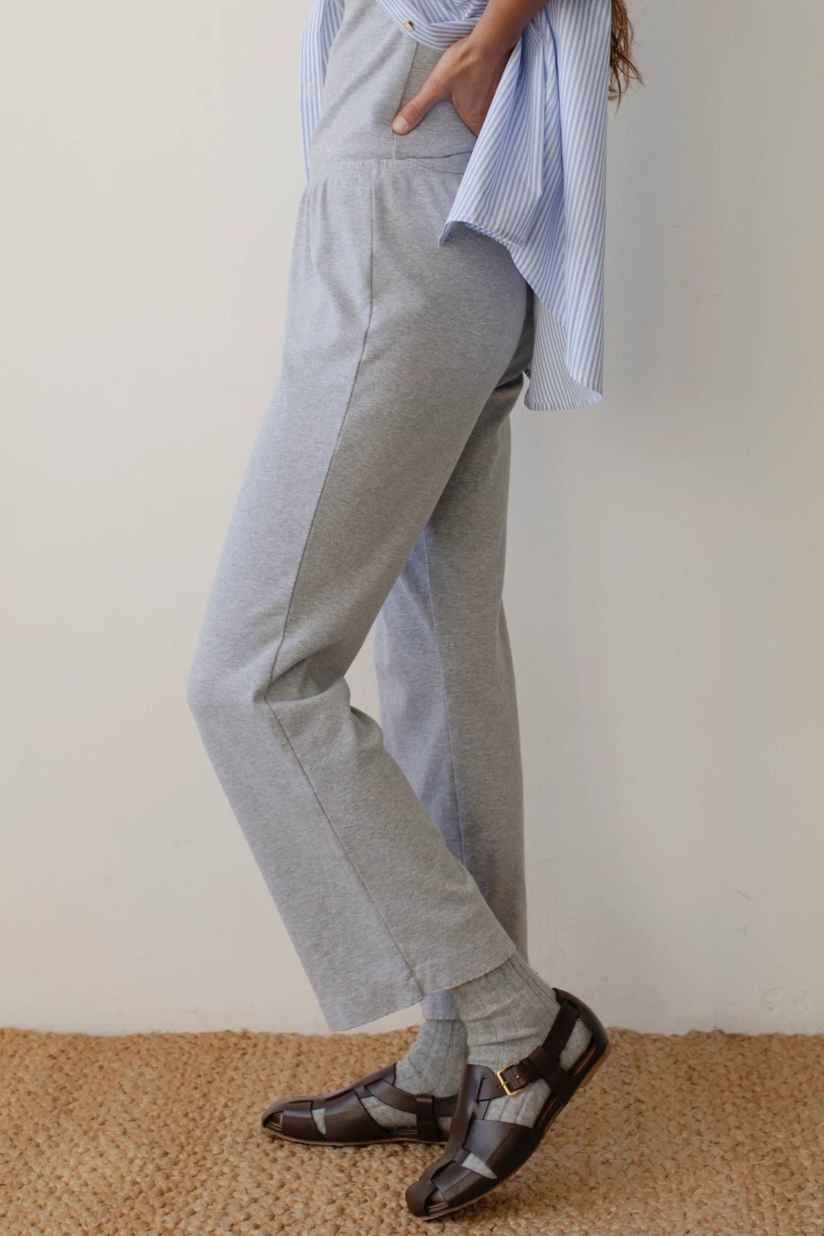 The person is wearing The Baby Rib Scallop Pant in light gray with a cropped fit by Donni, paired with a partially visible blue and white striped shirt, gray socks, and dark brown sandals. The casual outfit stands out against a simple background.