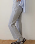 The person is wearing The Baby Rib Scallop Pant in light gray with a cropped fit by Donni, paired with a partially visible blue and white striped shirt, gray socks, and dark brown sandals. The casual outfit stands out against a simple background.