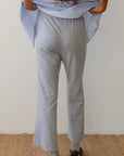 Rear view of a person wearing Donni's Baby Rib Scallop Pants in gray, paired with a blue and white striped shirt featuring scalloped edges, and black sandals worn over gray socks. They are standing on a woven rug against a white wall, their hands comfortably tucked into their shirt pockets.