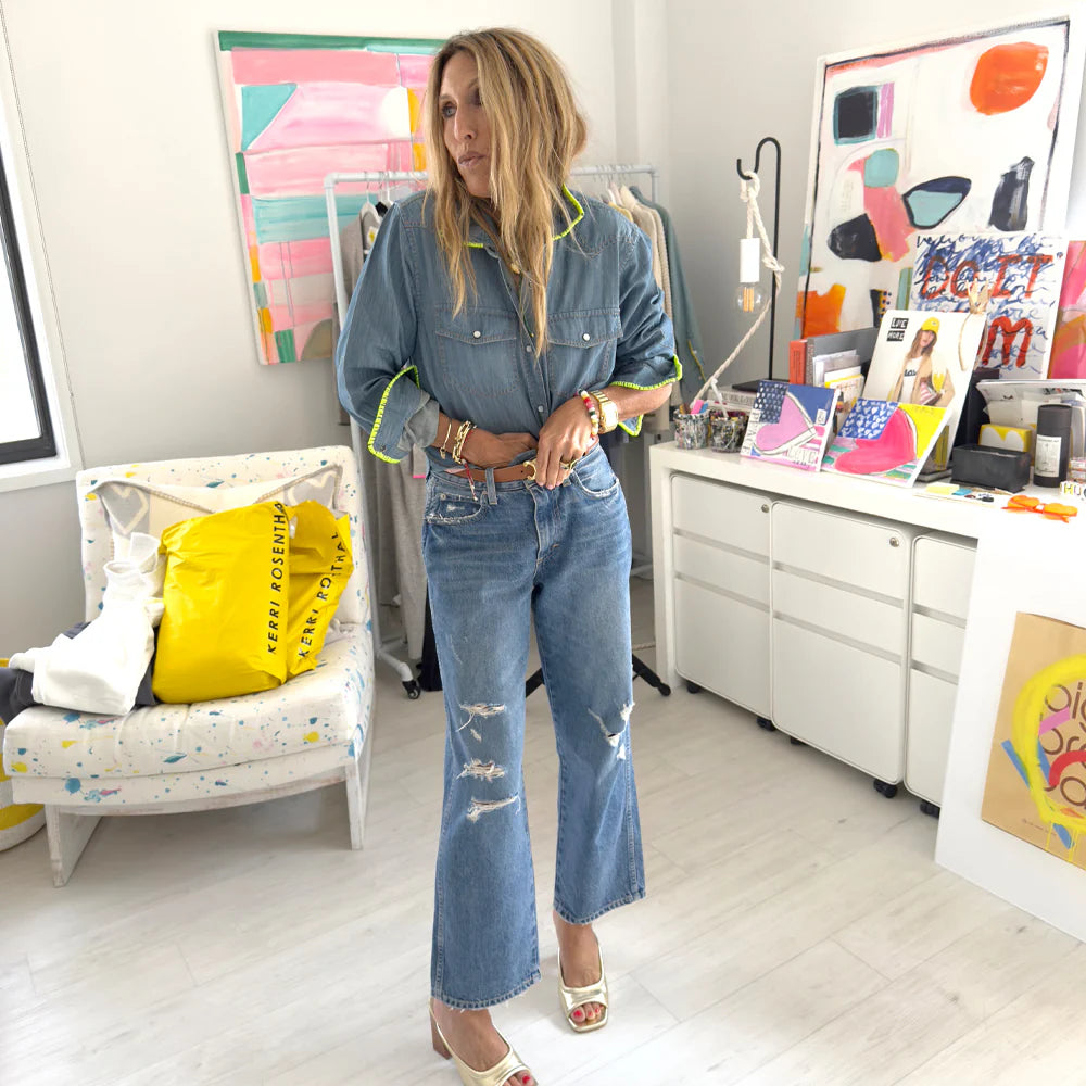 In a vibrant, art-filled room, a woman wears the Pia Denim Western Shirt by Kerri Rosenthal with distressed jeans and open-toe heels. She stands beside a painted chair with a yellow pillow, surrounded by paintings and art supplies.