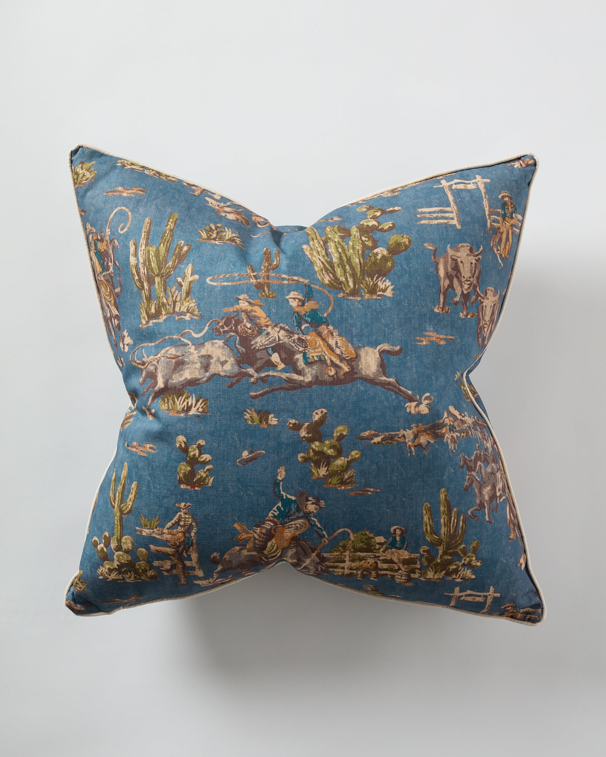 The Design Legacy Denim Cowboy Pillow 26x26 features a vintage Western print with cowboys, horses, cattle, cacti, and desert scenery on denim blue. Shown from above against a white surface.