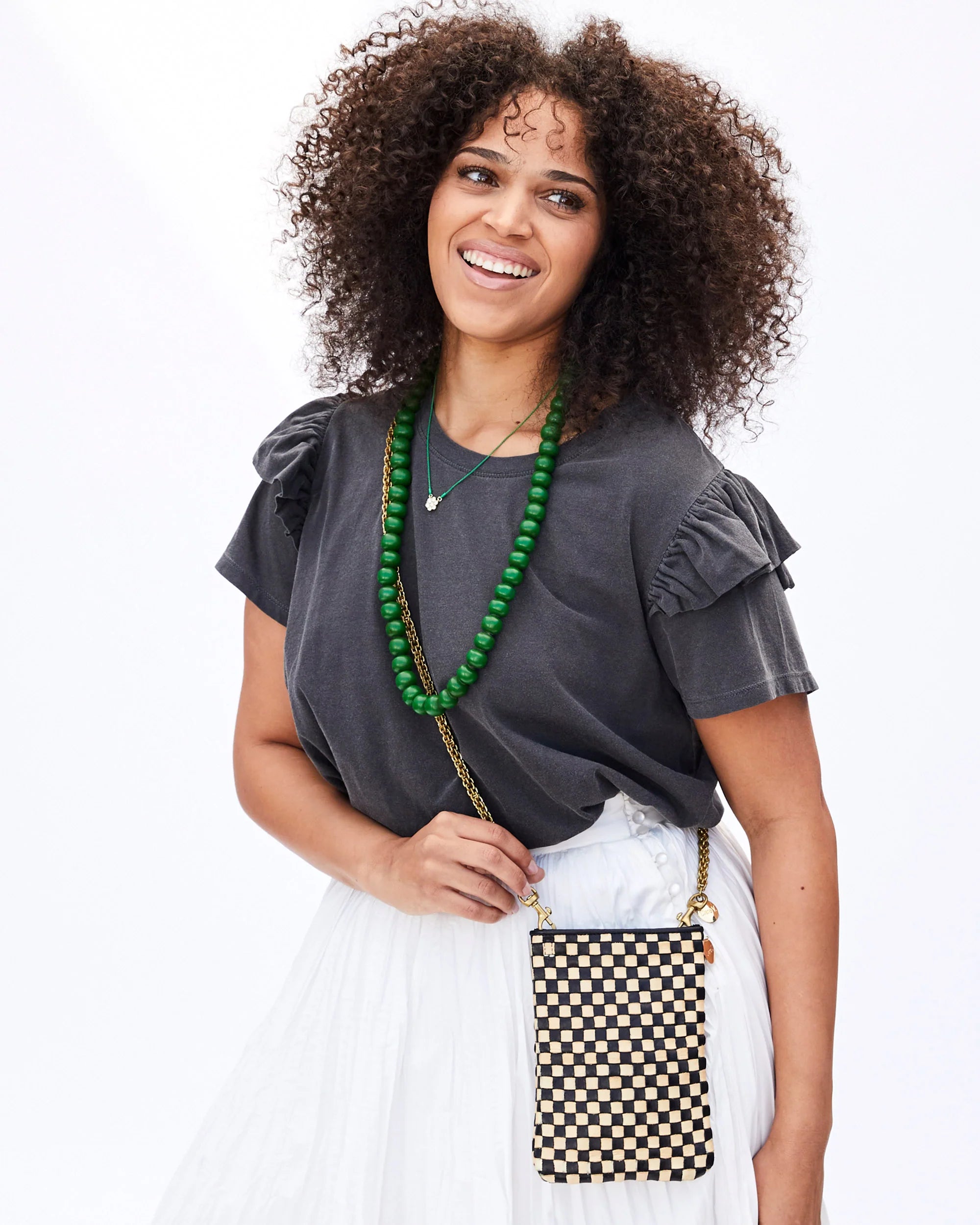 A person with curly hair smiles while posing against a white background. They wear a dark ruffled shirt, a green beaded necklace, and a white pleated skirt. They carry Poche by Clare Vivier, an everyday phone bag made of handwoven leather with a detachable crossbody strap.