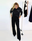 A person with long hair, dressed in a black sweater featuring white text and tailored Kerri Rosenthal Wide Leg Trouser Pants in black (XS), poses in a minimalist room. Beside them is a garment rack with clothes against a clean, white background.