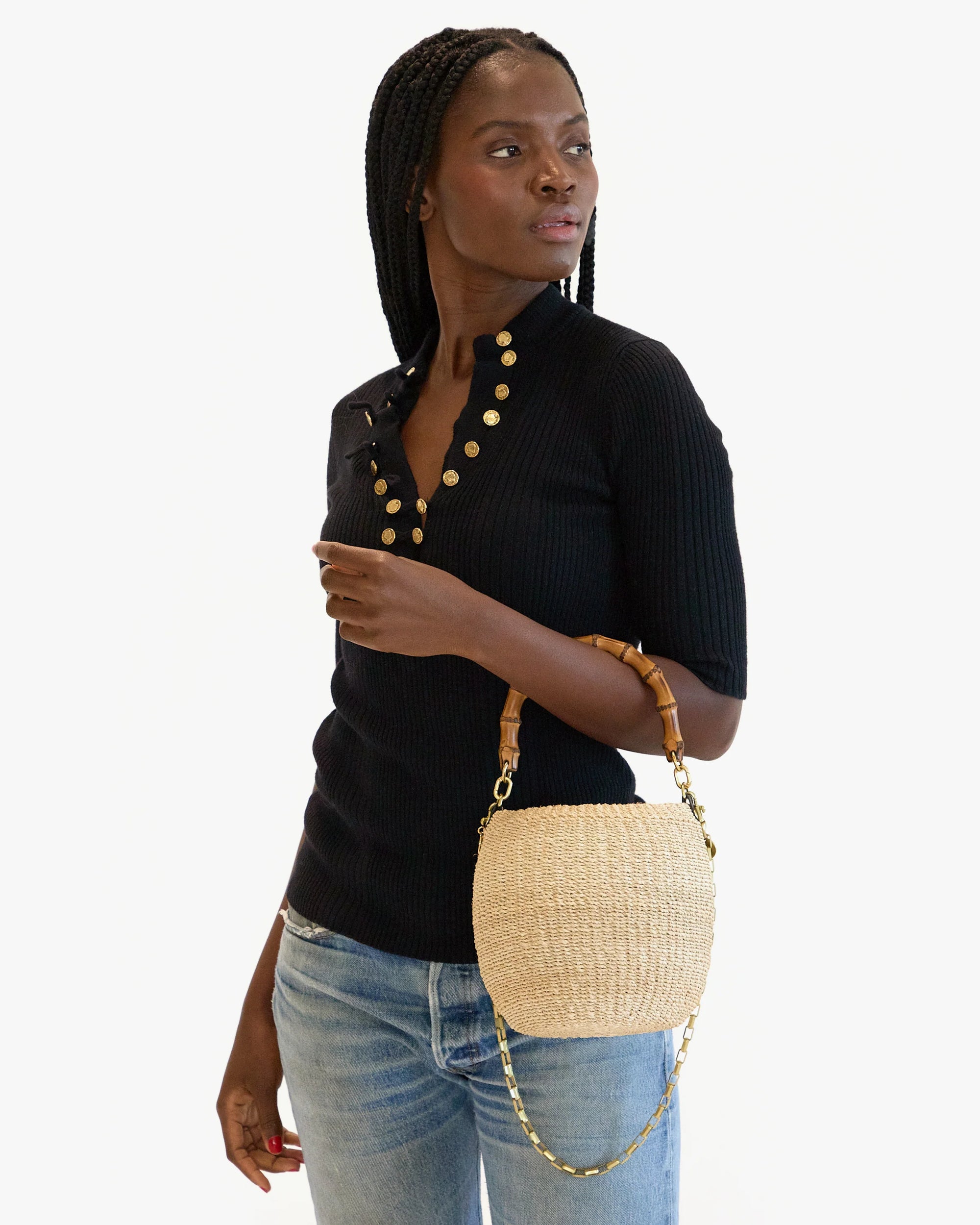 A person with long braided hair poses in a black ribbed top with gold buttons and blue jeans, holding Clare Vivier's Pot de Miel chic basket bag made from natural straw fibers, featuring an elegant bamboo handle. The background is white.