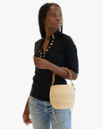 A person with long braided hair poses in a black ribbed top with gold buttons and blue jeans, holding Clare Vivier's Pot de Miel chic basket bag made from natural straw fibers, featuring an elegant bamboo handle. The background is white.