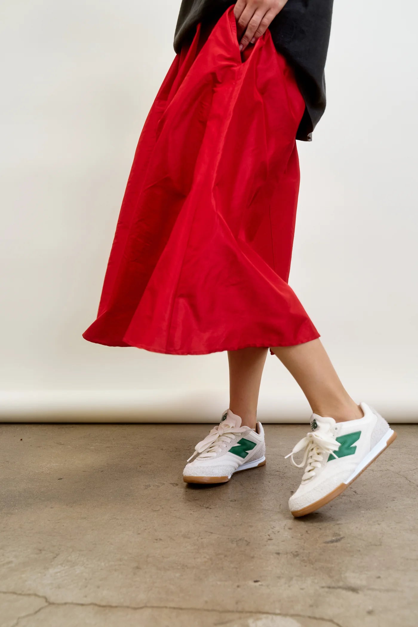 A person is seen from the waist down, walking on a concrete floor against a white backdrop. They're wearing LE SKIRT Silk Taffeta by Aquarius Cocktail, inspired by 1940s fashion in red with a flared design, along with white sneakers featuring green accents.