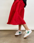 A person is seen from the waist down, walking on a concrete floor against a white backdrop. They're wearing LE SKIRT Silk Taffeta by Aquarius Cocktail, inspired by 1940s fashion in red with a flared design, along with white sneakers featuring green accents.