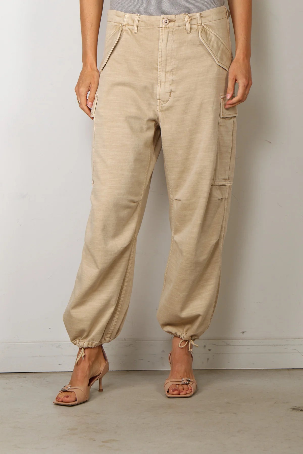 A person models the Denimist CROPPED CARGO pants—beige, relaxed-fit cargos with utility pockets—paired with open-toed heels and standing against a plain light-colored wall. Their arms rest naturally at their sides, and their head is out of frame.