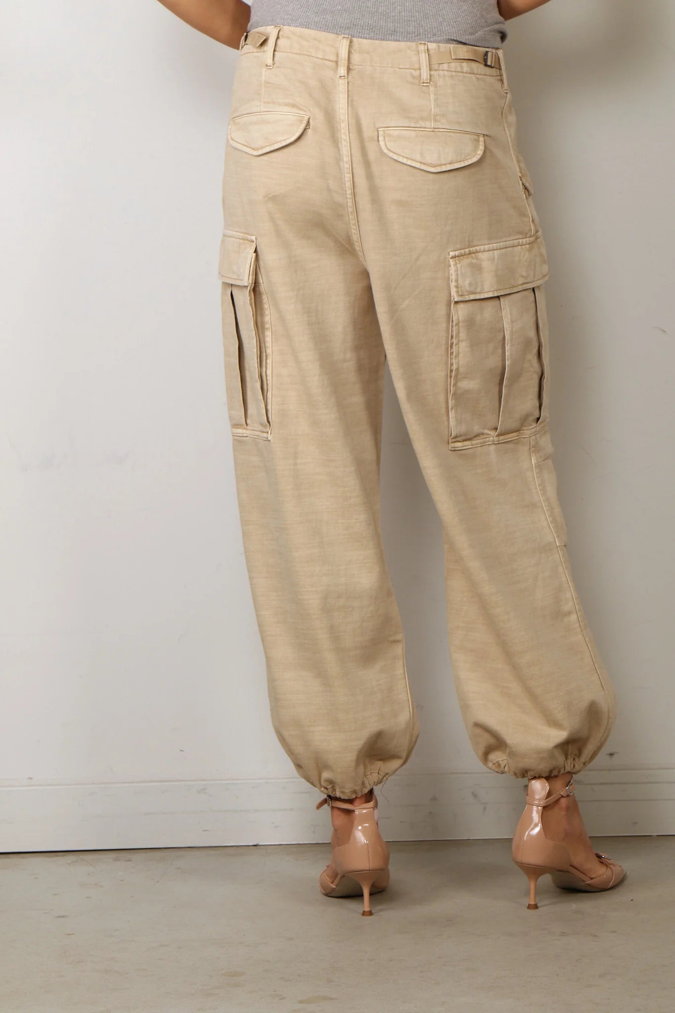A person wearing Denimist's CROPPED CARGO relaxed-fit beige pants featuring elastic cuffs and utility pockets, paired with high-heeled nude sandals, stands indoors facing a white wall.