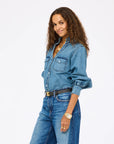 A woman with long curly hair smiles with one hand in her pocket, wearing the Kerri Rosenthal Puff Sleeve Shirt, blue jeans, a black belt, layered necklaces, and a gold watch against a plain white background.
