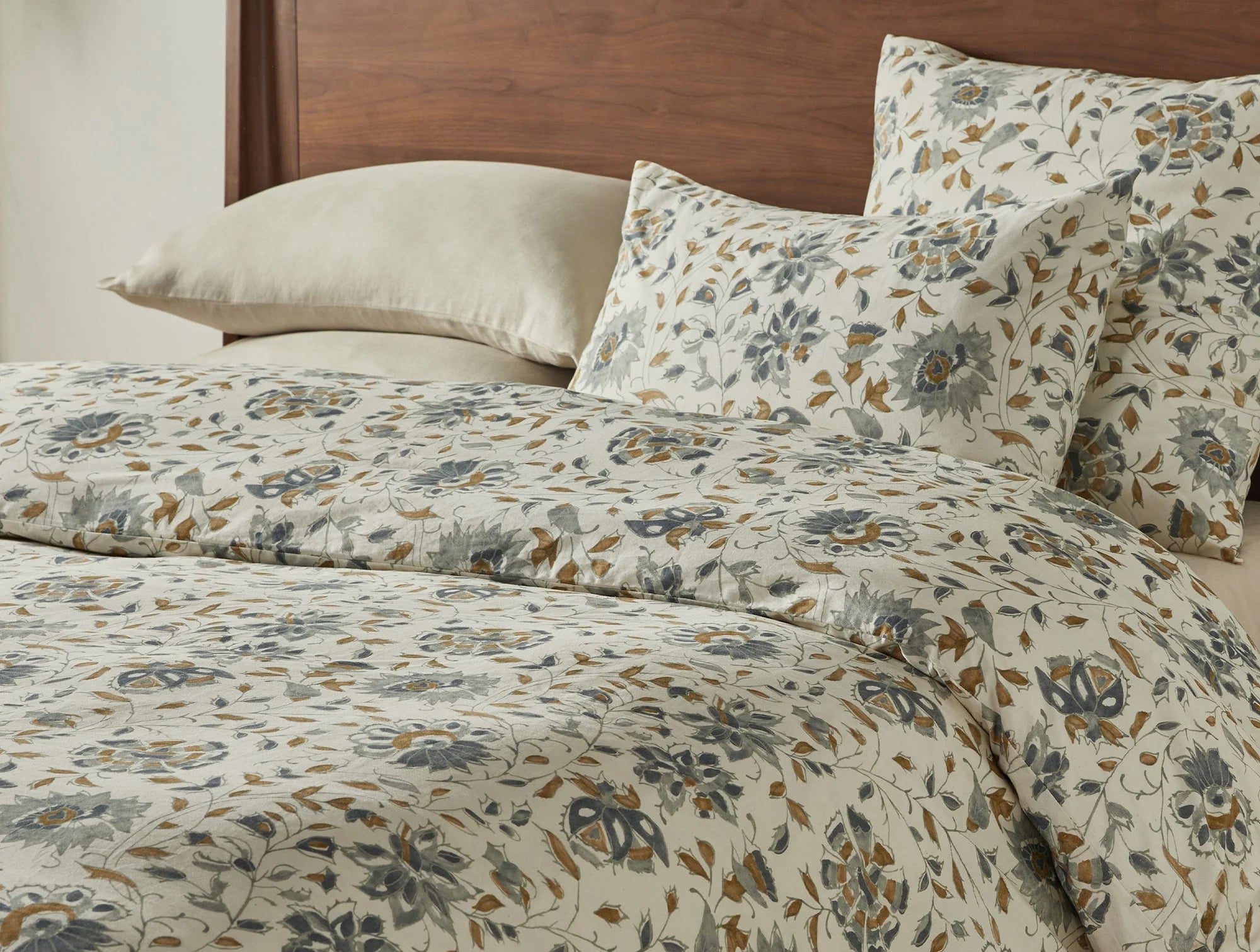 A bed with cream organic cotton sheets and pillows, plus the Coyuchi Inc Flora Org Print Duvet Undyed Lagoon King/CKing set—featuring a blue, gray, and tan botanical design—paired with a wooden headboard.