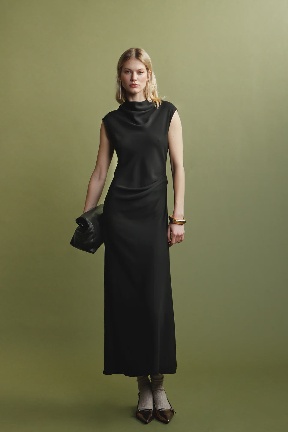 A woman with blonde hair and a neutral expression stands against an olive green background wearing the sleeveless black Naia Drape Dress by Psophia, brown shoes with light socks, and holding a black clutch.