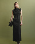 A woman with blonde hair and a neutral expression stands against an olive green background wearing the sleeveless black Naia Drape Dress by Psophia, brown shoes with light socks, and holding a black clutch.