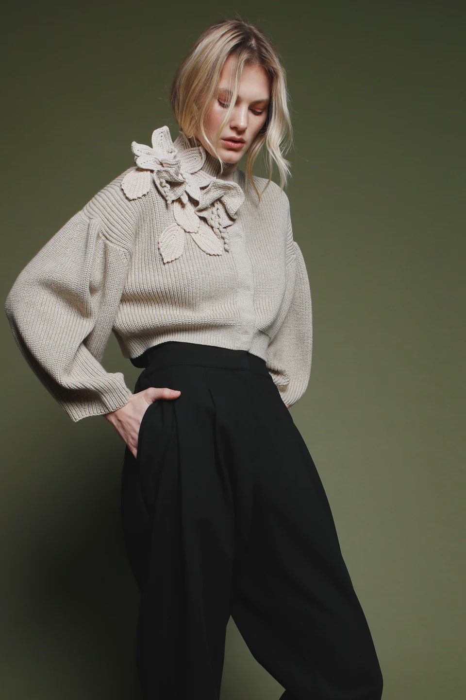 A woman with blonde hair poses against a green background, wearing the Psophia Funnel Neck Cardigan in light beige with floral shoulder detail and high-waisted black wide-leg pants, looking down with one hand in her pocket.