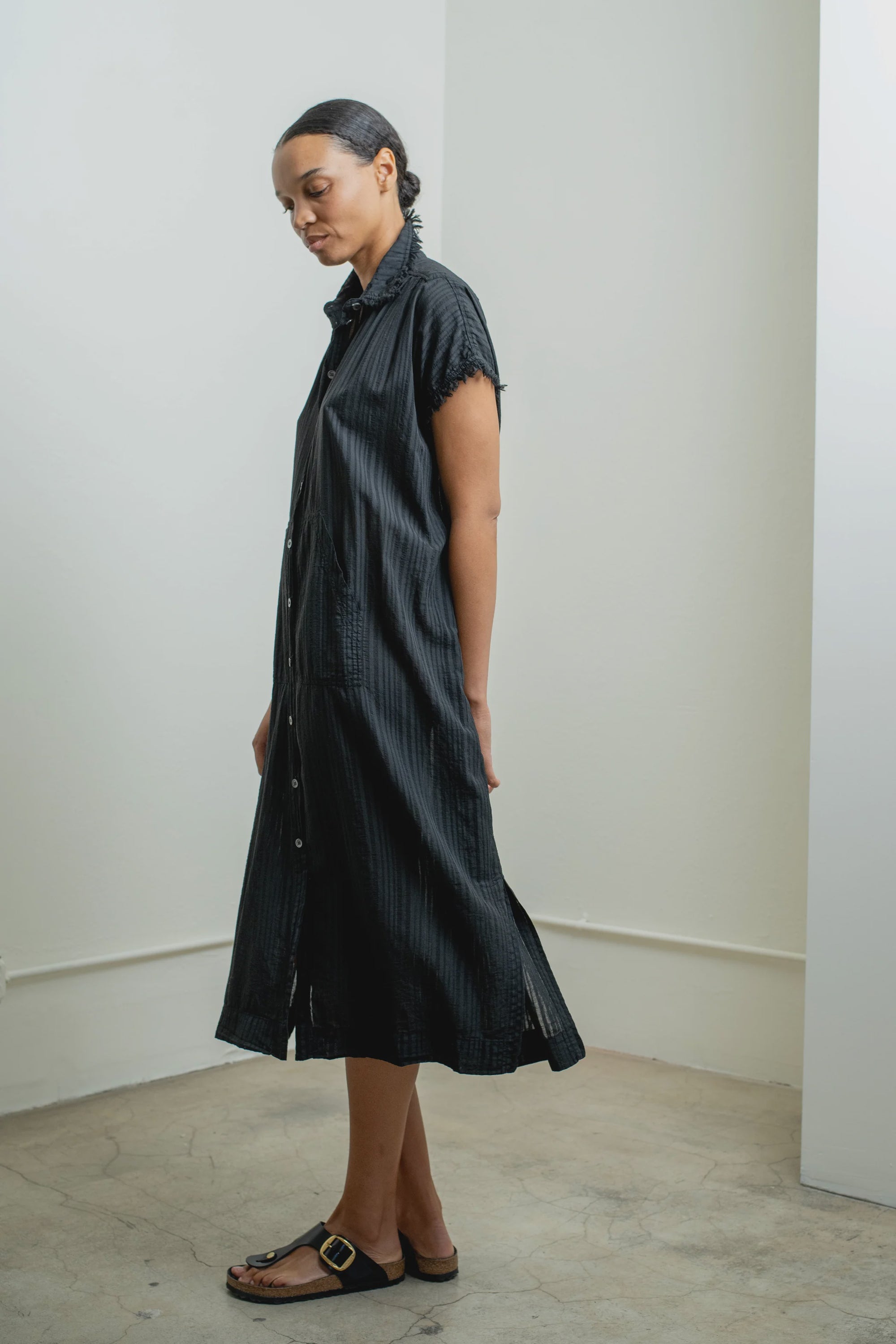 A woman stands sideways in a minimalist room, wearing the Raquel Allegra SS Caftan in black and matching sandals. Her hair is tied back, and she gazes down with a calm expression.
