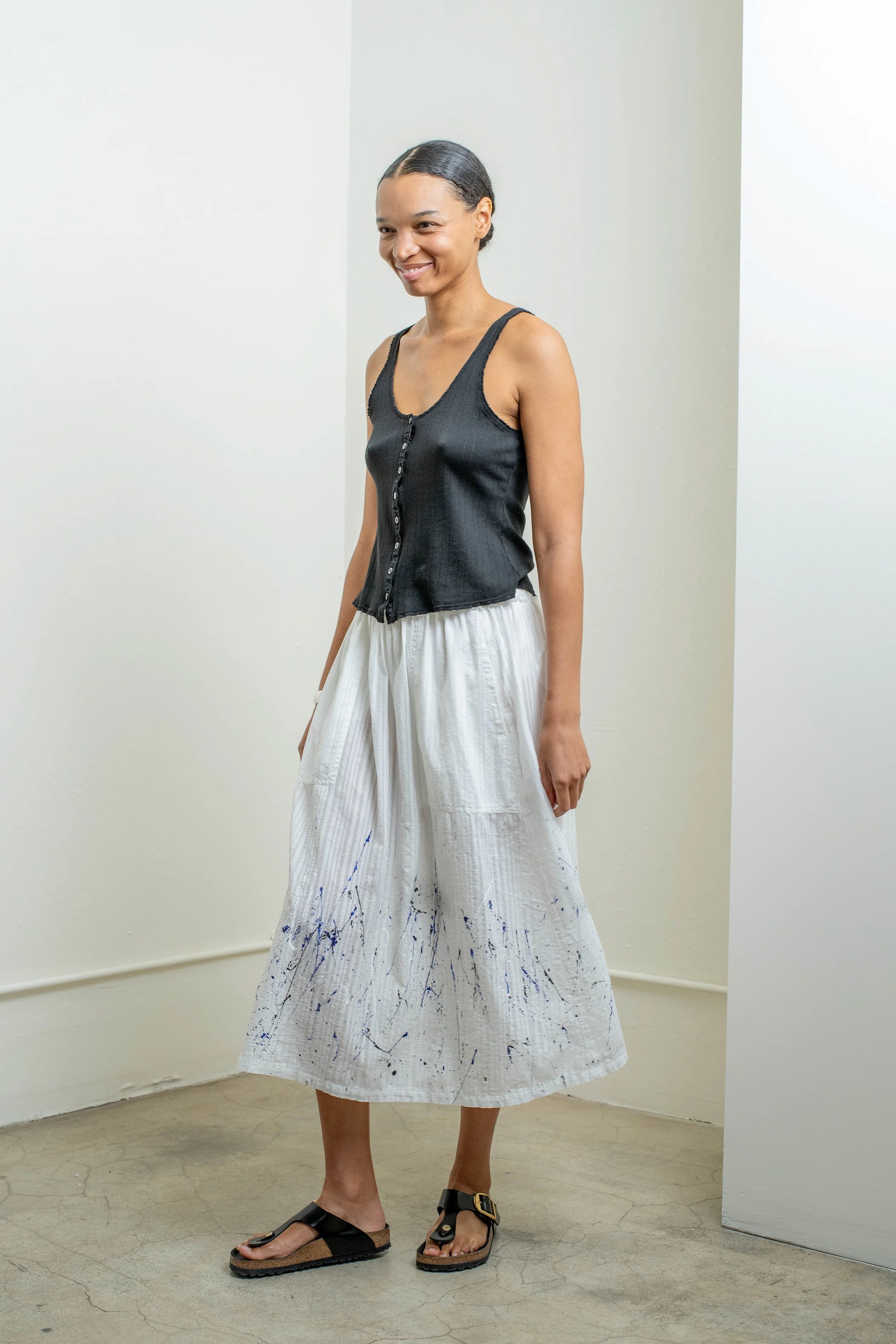 A person stands indoors by a white wall, smiling and wearing a dark sleeveless top, black sandals, and the Raquel Allegra Day Skirt—a long white skirt featuring a blue hand-painted print.