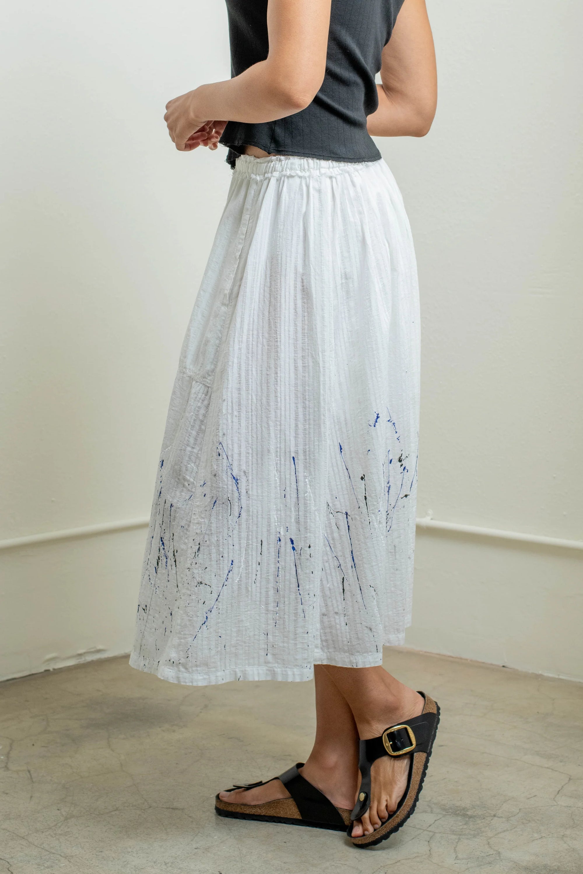 A person wears a black sleeveless top, black sandals, and a Raquel Allegra Day Skirt—a white cotton piece with subtle blue hand-painted splatters—while standing indoors on a light-colored floor.