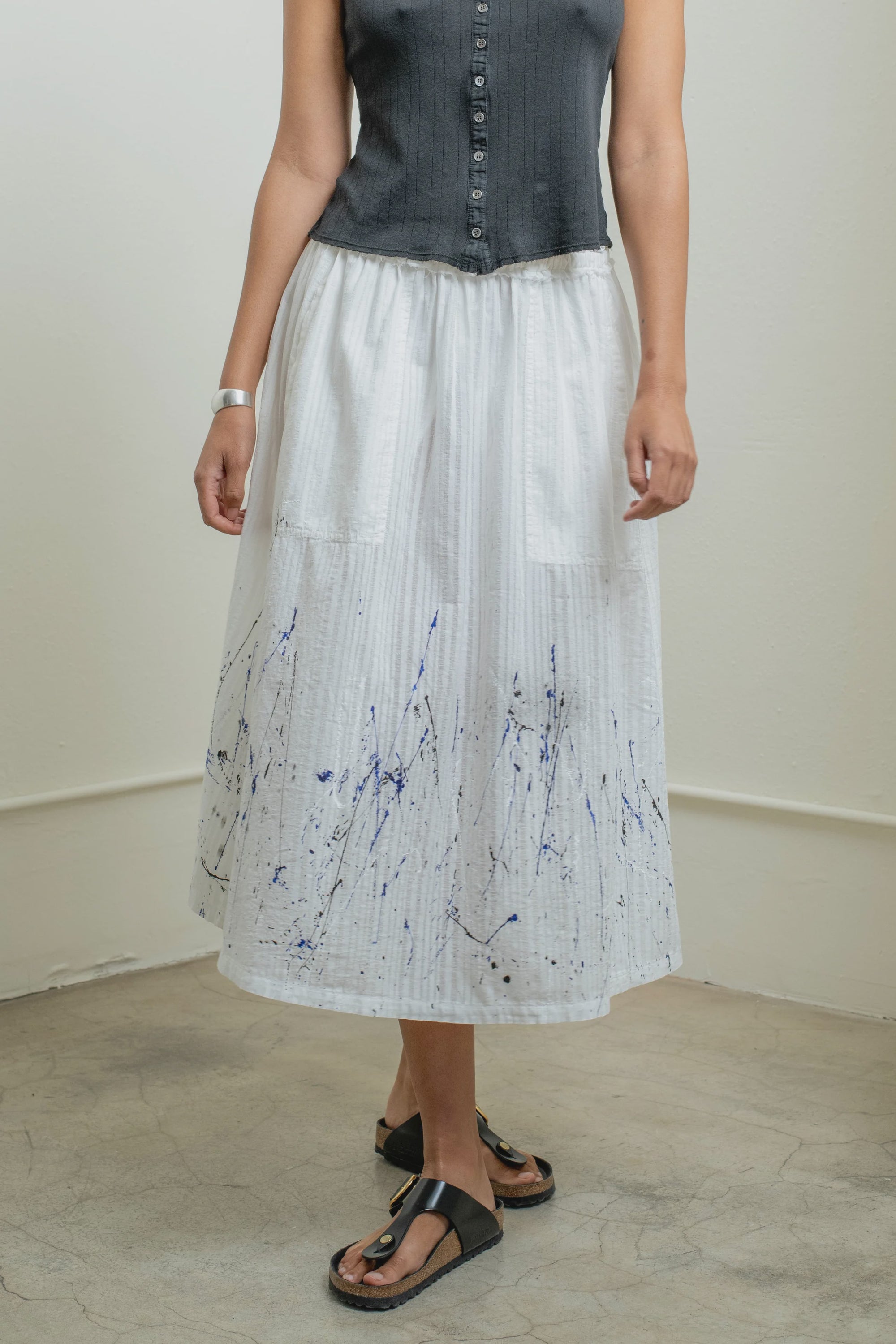 A person stands indoors on a concrete floor wearing a sleeveless black button-up top, Raquel Allegra Day Skirt with blue splatter print near the hem, black sandals, and a silver bracelet.