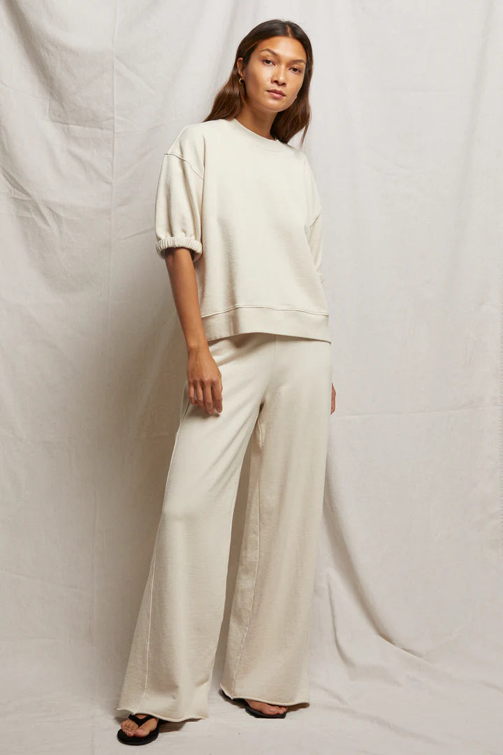 Against a light fabric backdrop, a person with long hair wears the Rebecca 3/4 Puff Sleeve top by Perfectwhitetee in cream. The oversized fit paired with matching wide-leg pants is crafted from cozy French terry cotton. They look neutrally at the camera.