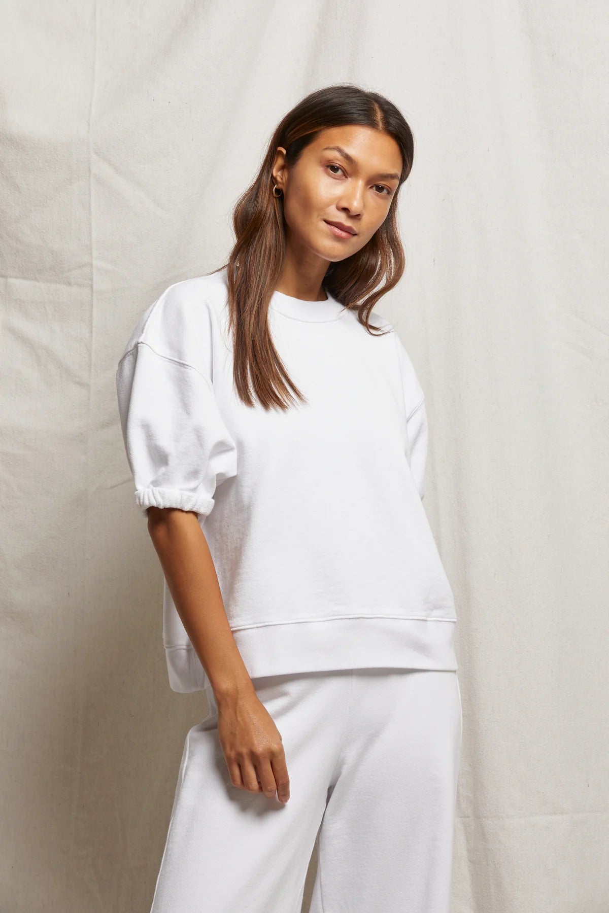 A woman with long brown hair wears the Rebecca SS Crew, a soft cotton white sweatshirt from Perfectwhitetee, paired with matching pants. She stands in front of a light beige fabric backdrop, looking relaxed with her left hand by her side.
