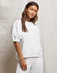A woman with long brown hair wears the Rebecca SS Crew, a soft cotton white sweatshirt from Perfectwhitetee, paired with matching pants. She stands in front of a light beige fabric backdrop, looking relaxed with her left hand by her side.