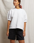 A woman stands against a light fabric backdrop wearing the Perfectwhitetee Rebecca SS Crew, black denim shorts, and a pink hair scarf. She looks to the side with a relaxed posture.
