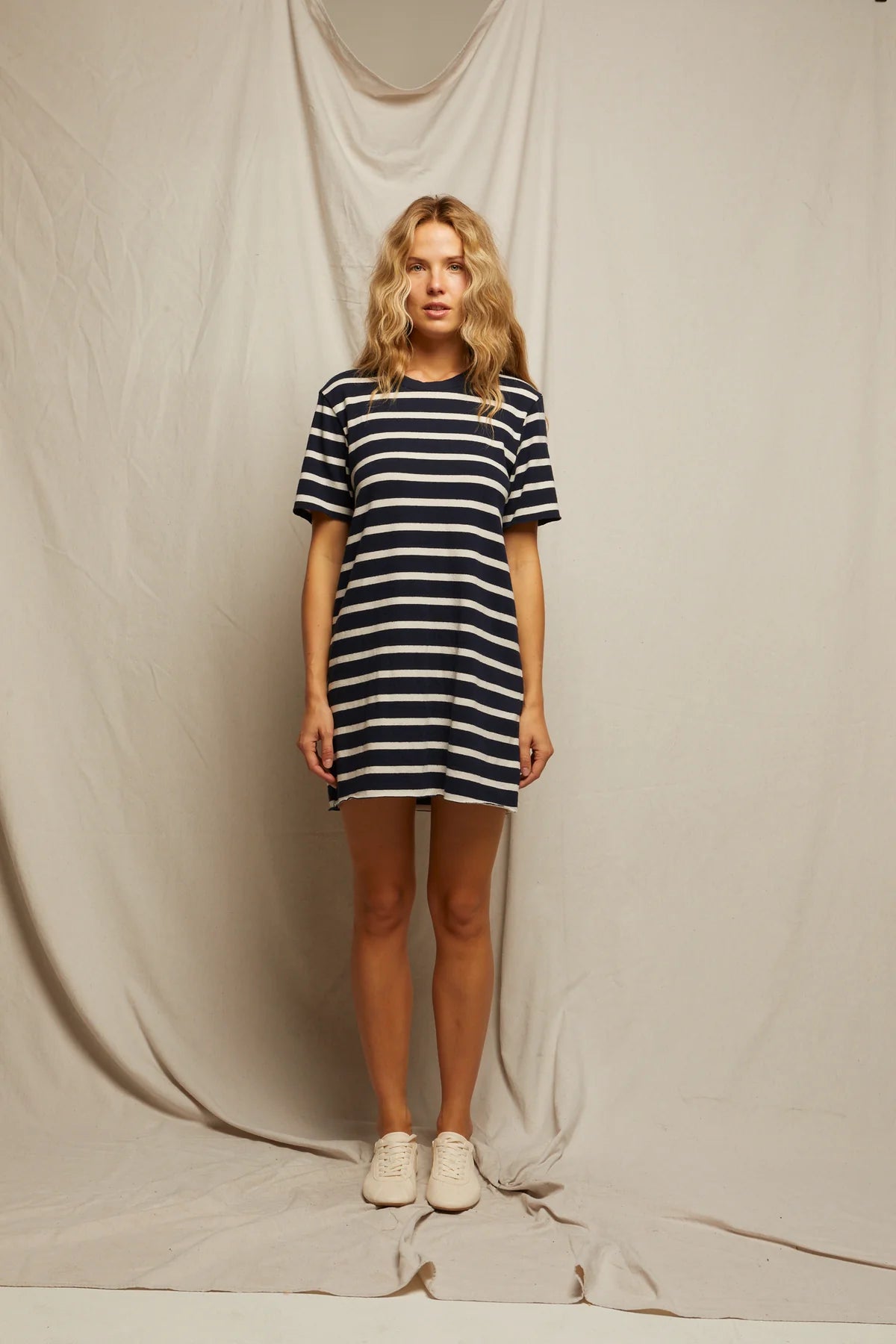 A woman faces forward in front of a beige fabric backdrop, wearing the Perfectwhitetee Rhett SS Tee Dress—a relaxed, black and white striped dress—paired with light sneakers and a neutral expression.