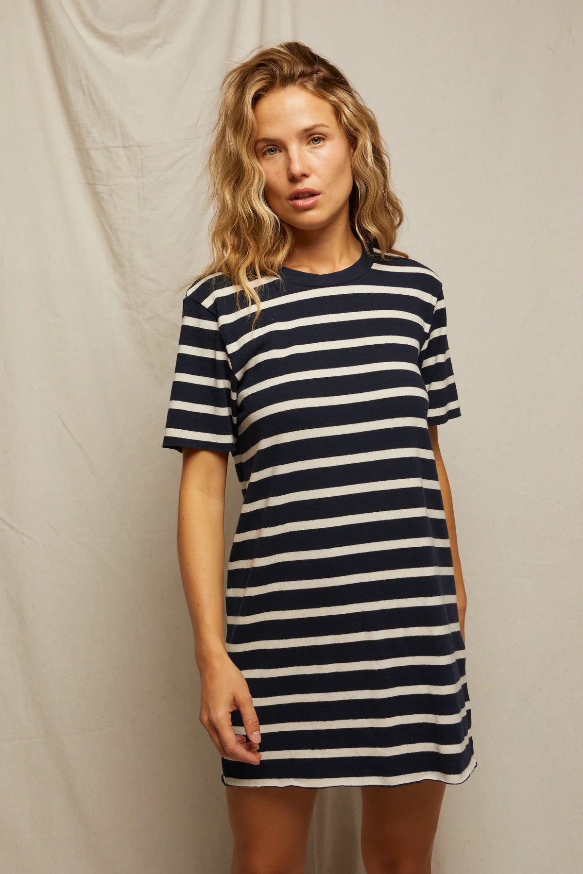 A striped beauty with wavy blonde hair models the Rhett SS Tee Dress by Perfectwhitetee—a short-sleeved, navy blue and white t-shirt dress—against a plain light backdrop.