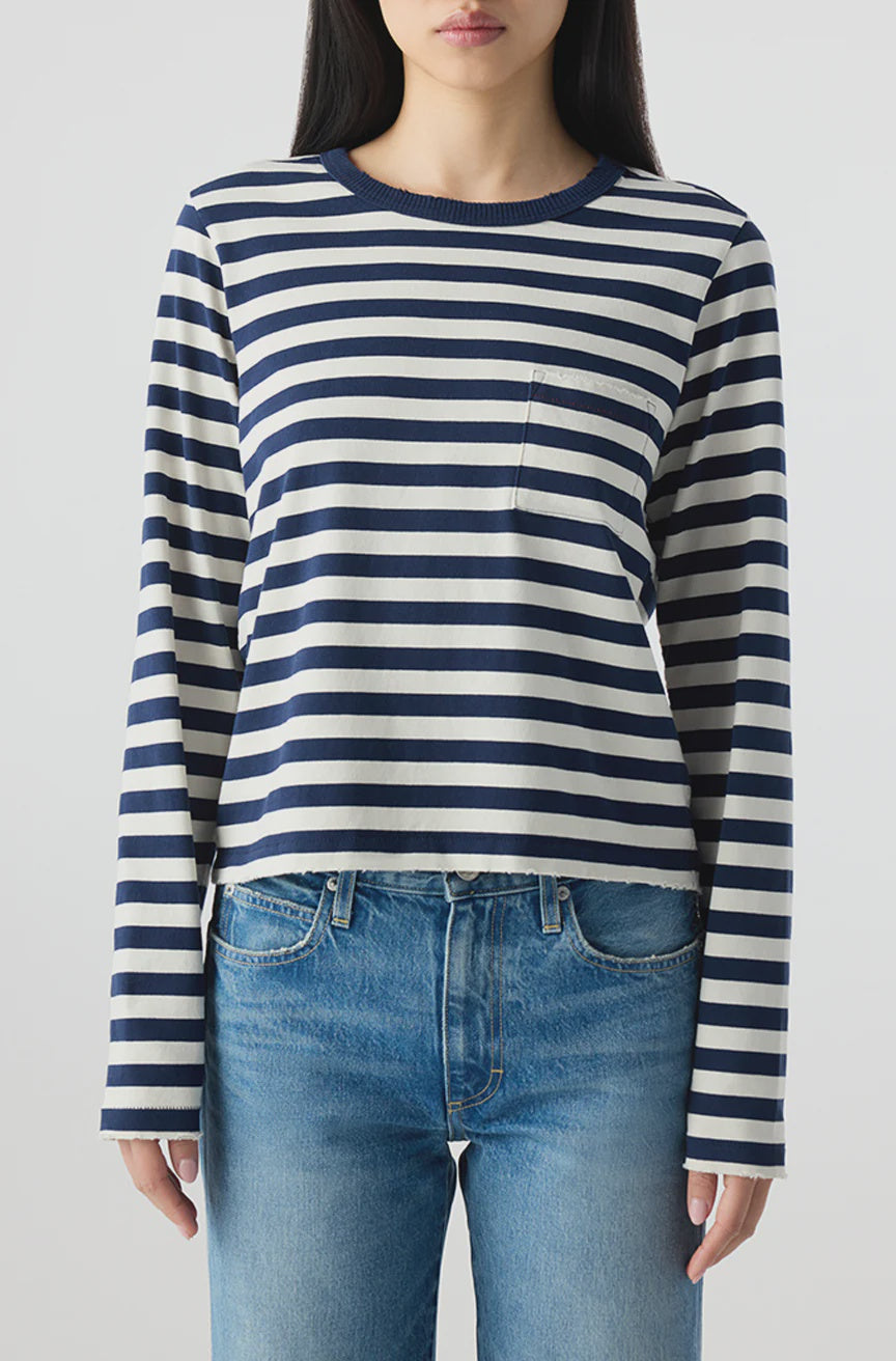 A person wears the Brigitte LS Tee by AMO Denim—navy and white stripes with a chest pocket—paired with relaxed fit blue jeans, against a plain light background. The casual Los Angeles-made look features the face partially out of frame.