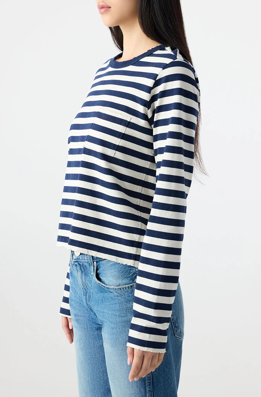 A woman with long dark hair models the AMO Denim Brigitte LS Tee, a relaxed-fit navy and white striped top, paired with blue jeans and shown in a three-quarter side view against a plain white background.