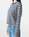 A woman with long dark hair models the AMO Denim Brigitte LS Tee, a relaxed-fit navy and white striped top, paired with blue jeans and shown in a three-quarter side view against a plain white background.