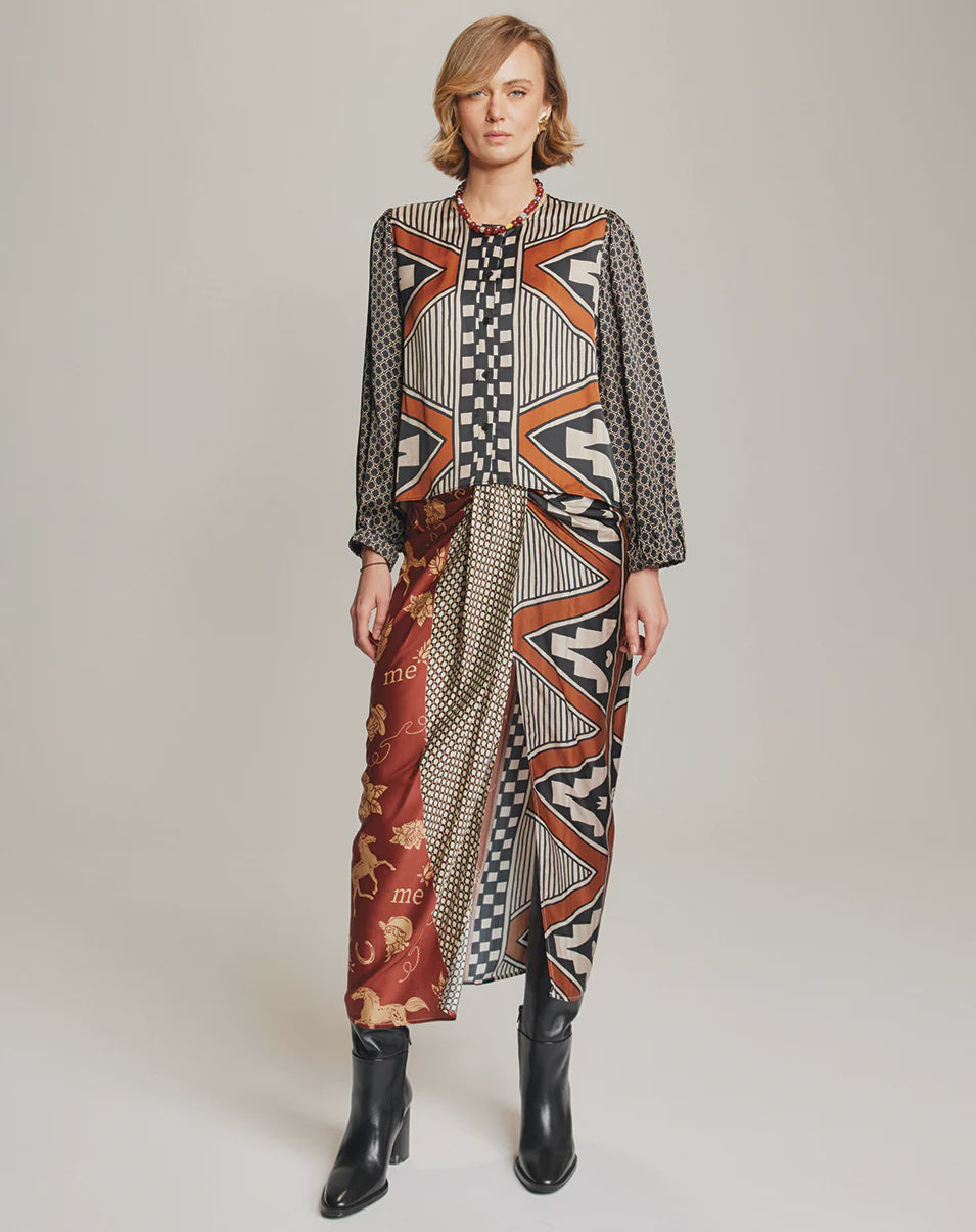 A woman with short blonde hair faces forward, wearing the Me369 Avia Round Neck Shirt and a skirt with bold geometric patterns in black, white, red, and gold. She completes the look with black ankle boots.