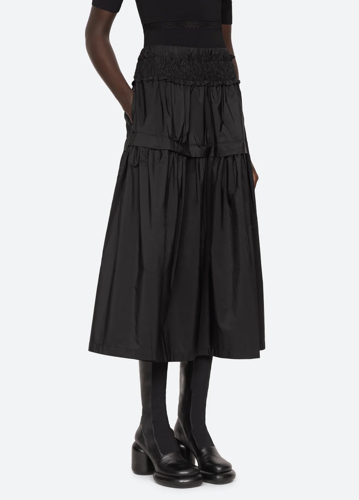 A person wearing the black Belen Taffeta Smocked Tiered Skirt by Sea New York, paired with a matching black top and platform shoes. The high-waisted design and midi silhouette are accentuated by its layered and pleated structure, adding texture to the ensemble. The background is plain and light-colored.