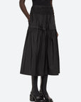 A person wearing the black Belen Taffeta Smocked Tiered Skirt by Sea New York, paired with a matching black top and platform shoes. The high-waisted design and midi silhouette are accentuated by its layered and pleated structure, adding texture to the ensemble. The background is plain and light-colored.