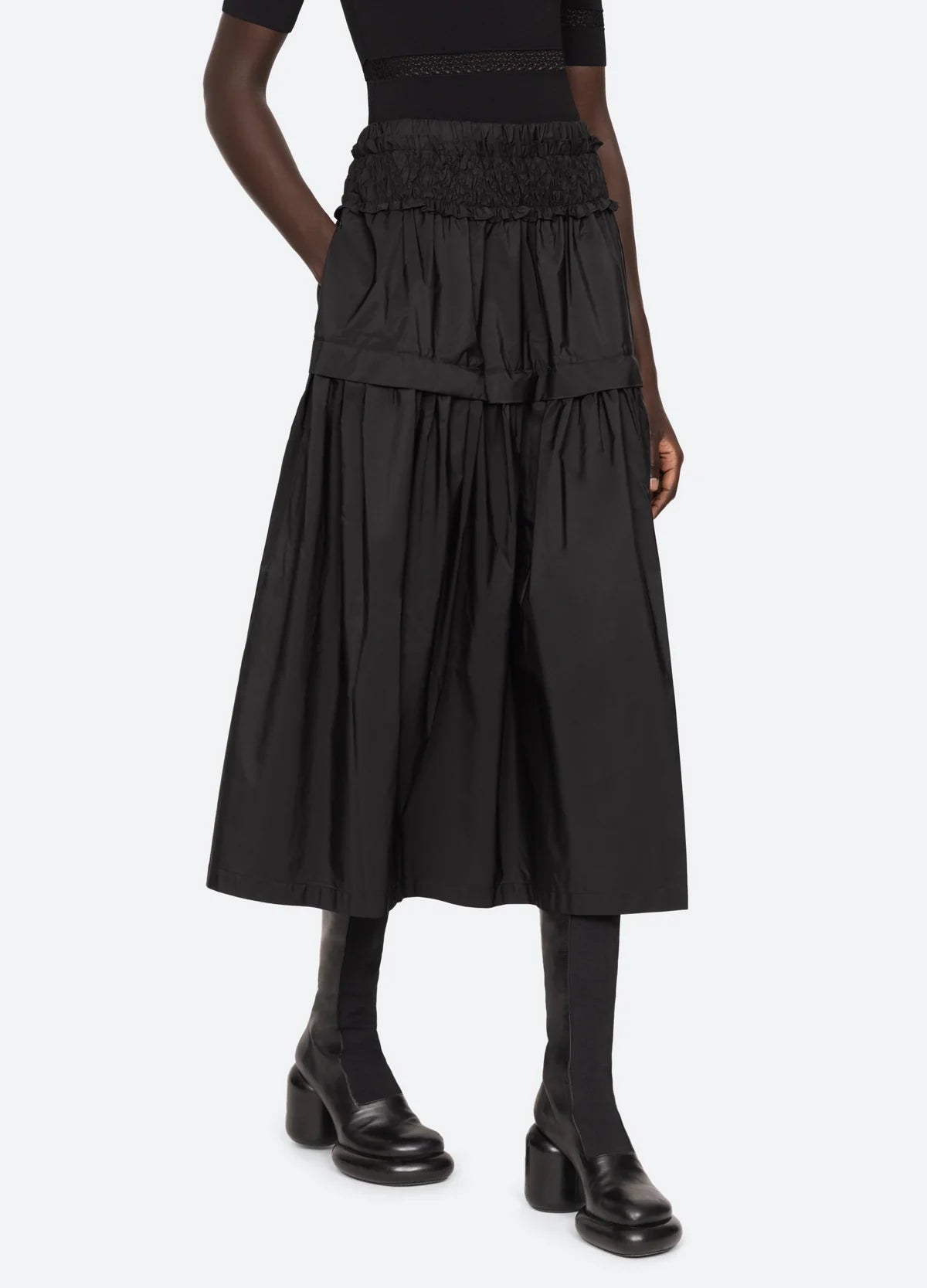 A person stands against a white background, wearing the Sea New York Belen Taffeta Smocked Tiered Skirt in a textured black fabric, paired with black heeled boots featuring a chunky heel.