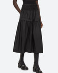 A person stands against a white background, wearing the Sea New York Belen Taffeta Smocked Tiered Skirt in a textured black fabric, paired with black heeled boots featuring a chunky heel.