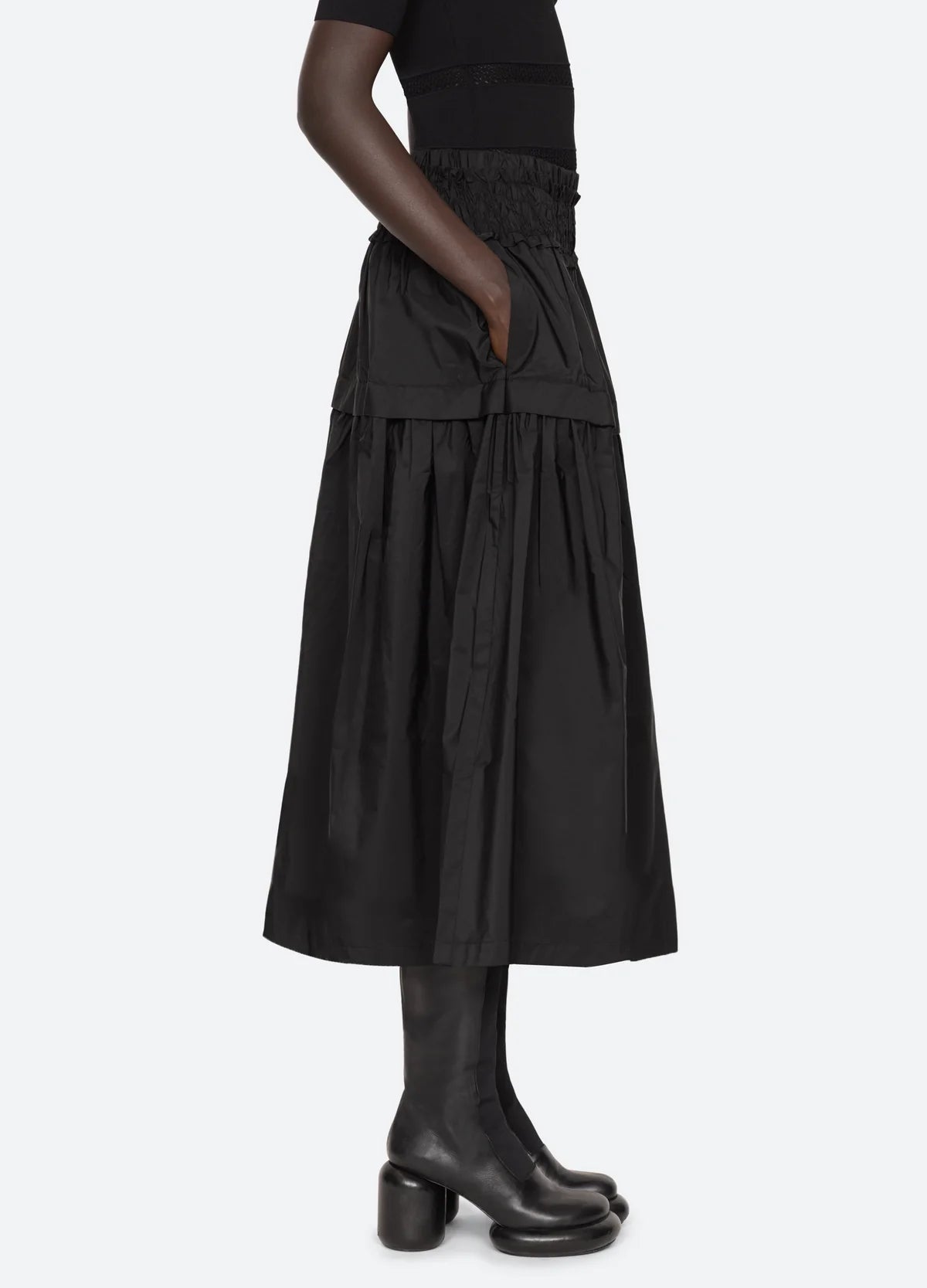 A person wearing the Belen Taffeta Smocked Tiered Skirt by Sea New York, paired with black tall boots, stands sideways against a plain light background.