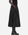 A person wearing the Belen Taffeta Smocked Tiered Skirt by Sea New York, paired with black tall boots, stands sideways against a plain light background.